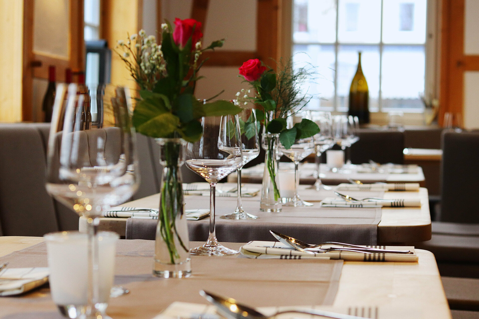 Ein gedeckter Tisch in einem Restaurant mit sechs Weingläsern, zwei Vasen mit roten Rosen und Silberbesteck, mit einem Fenster und Holzbalken im Hintergrund.