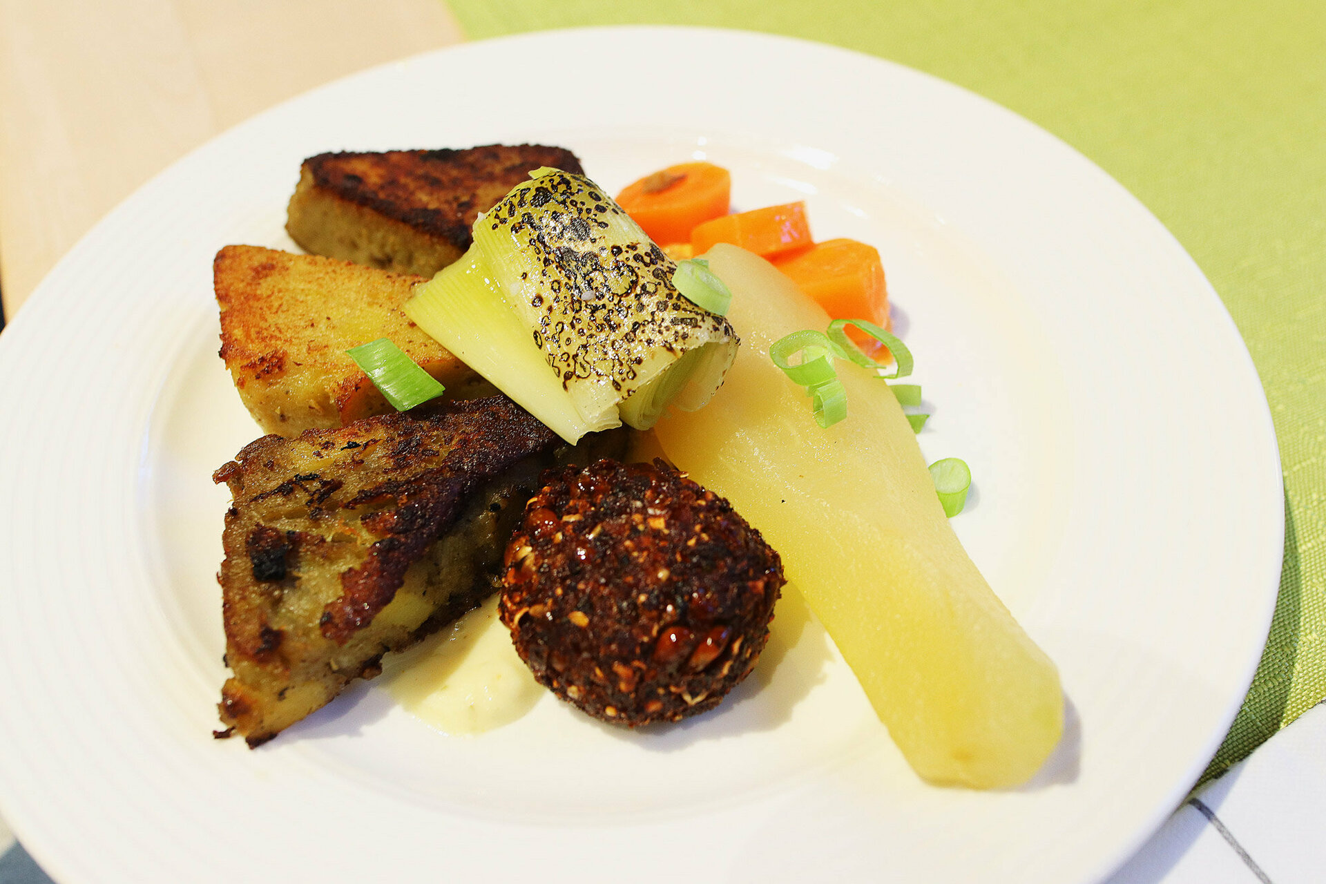 Auf einem weißen Teller liegt eine bunte Auswahl an Speisen, darunter ein Stück gegrilltes Fleisch, eine gelbe Birne, eine dunkelbraune Frikadelle und orangefarbene Karotten, garniert mit grünen Zwiebeln.