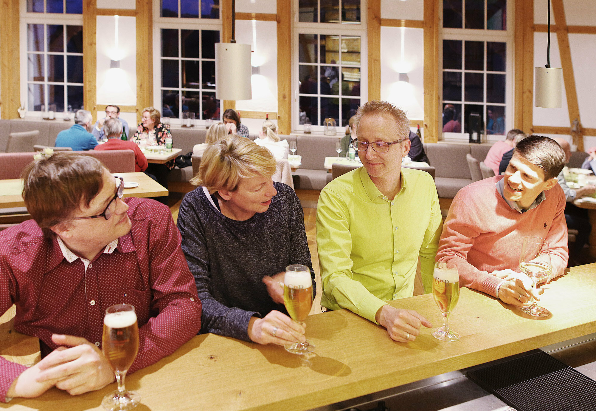 Vier Personen unterhalten sich an einer hölzernen Bar in einem gemütlichen Restaurant, umgeben von Tischen, Stühlen und einem warmen Ambiente.