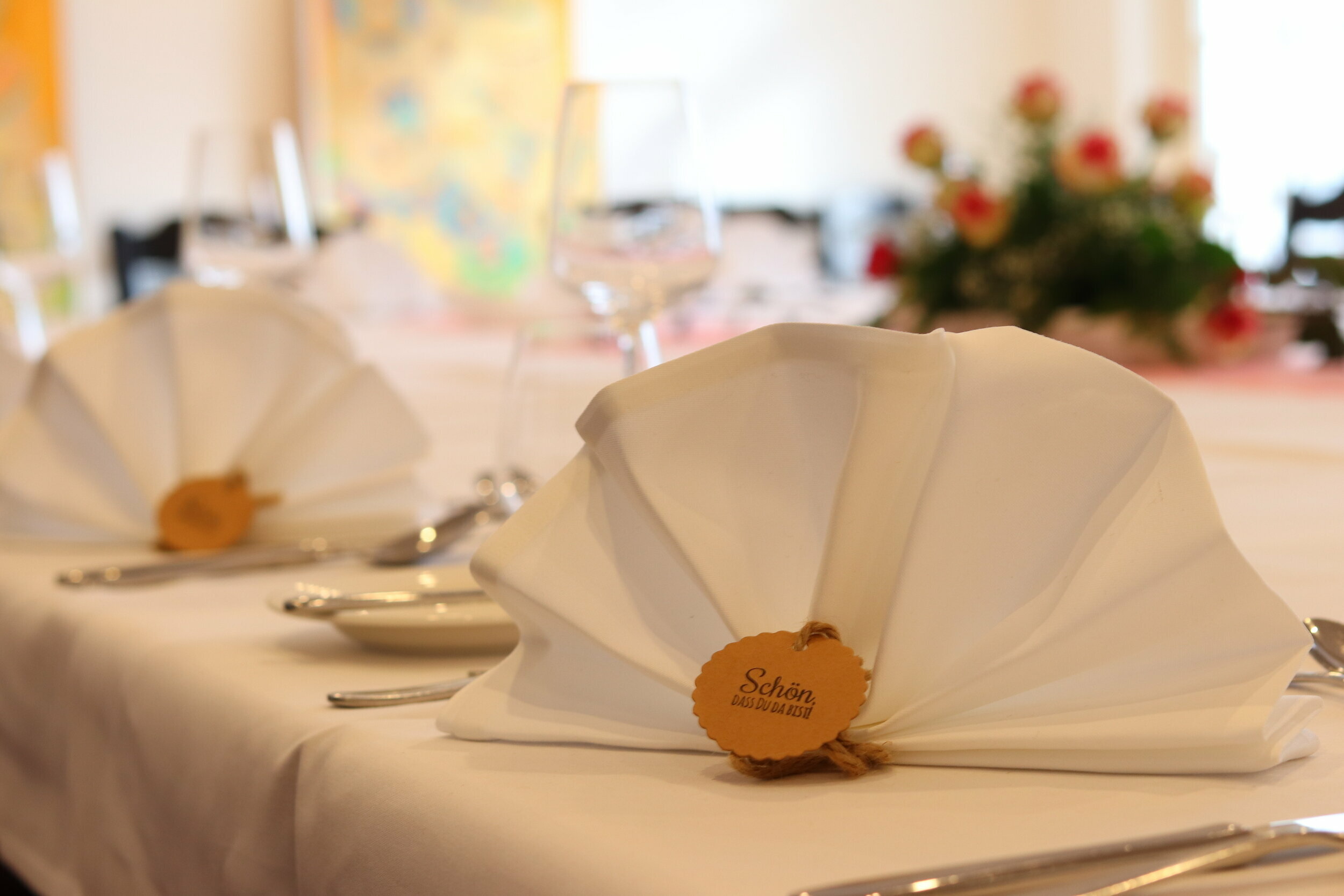 Ein Tisch für eine formelle Veranstaltung ist mit weißer Tischdecke, Silberbesteck und einem Mittelstück aus roten Rosen gedeckt, auf dem ein kleines Holzschild mit der Aufschrift &quot;Schön, dass das ist&quot; steht.
